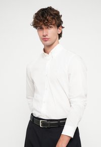 Bílá košile s knoflíky, strukturovaným límcem, dlouhými rukávy a malým logem na levé straně hrudi, doplněná černým pásem a kalhotami.