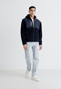 Zwart fleecejack voor heren met marineblauwe panelen, ritssluiting, capuchon met trekkoord en rode accenten. Draagt met lichtblauwe jeans en witte sneakers.