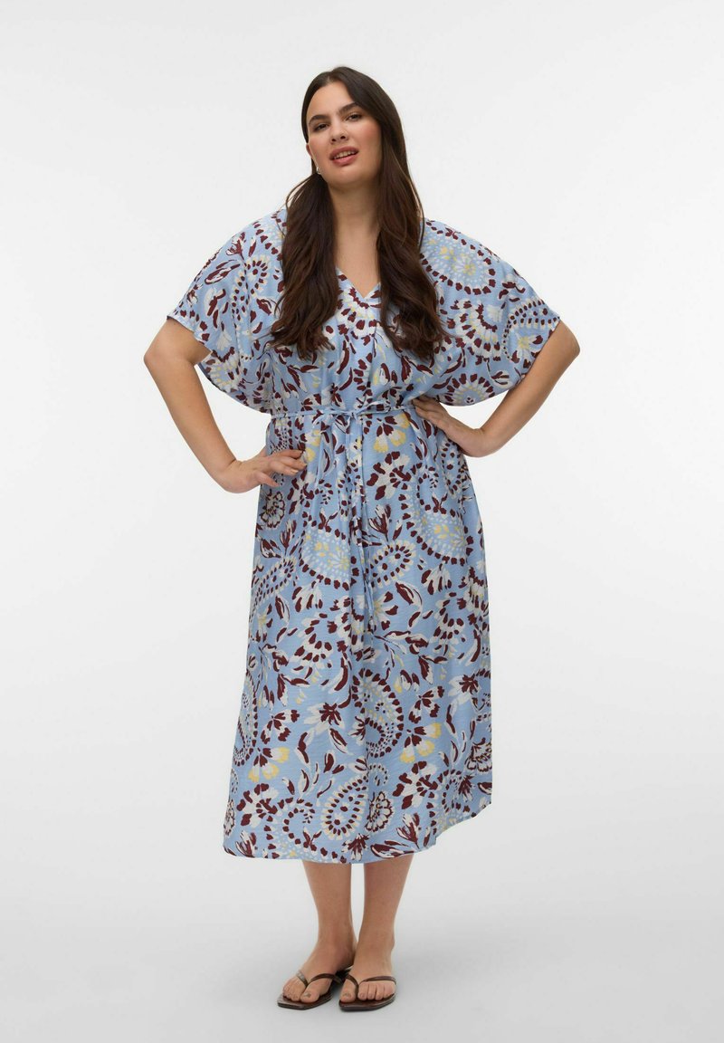 Femme aux longs cheveux noirs portant une robe midi bleue à motifs avec des manches courtes et des sandales noires, debout les mains sur les hanches.