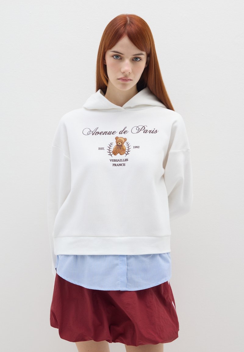 Witte hoodie met geborduurde tekst en berenontwerp. Draagt over een lichtblauw gestreept shirt en rode shorts. Soepele textuur.