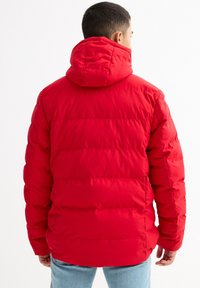 Hombre con chaqueta acolchada roja brillante con capucha, de espaldas, combinado con jeans azul claro, sobre fondo blanco liso.