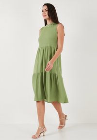 Green, sleeveless dress with a round neckline and tiered design. Lightweight, textured fabric. Paired with light strappy sandals.