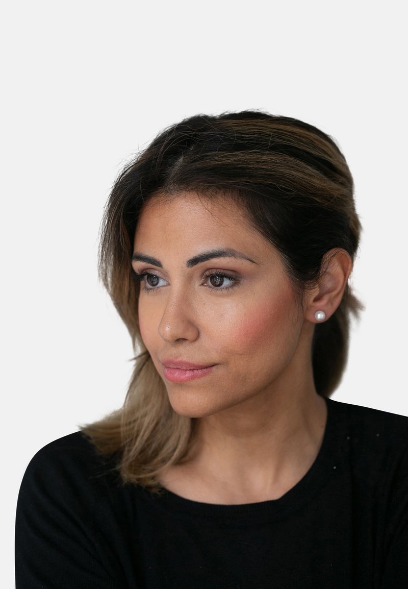 Porträt einer Frau mit schulterlangen braunen Haaren, die ein schwarzes Oberteil und Perlenohrringe trägt, vor einem neutralen Hintergrund und mit natürlichem Make-up.