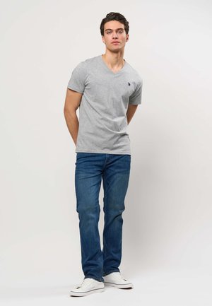 Mladý muž stojí s rukami za chrbtom, má na sebe sivé tričko s výstrihom do V, modré džínsy a biele tenisky na jednotnom svetlom pozadí.