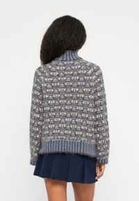 Person med langt mørkt hår iført en blå, grå og hvid stribet fuzzy sweater og en marineblå plisseret nederdel, der står med ryggen til kameraet.