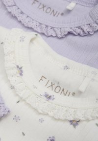 Deux chemises à col rond : l'une lavande en côtelé, l'autre blanche avec un imprimé floral violet et un bord en dentelle. Étiquettes visibles, détails clairs.
