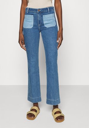 Flared blue jeans with contrasting light blue back pockets, high-waisted design, and a raw hem. Paired with tan slip-on sandals.