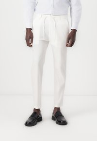 Pantalons blancs à cordon de serrage en tissu léger, avec une coupe fuselée et une texture lisse. Portés avec des mocassins noirs.