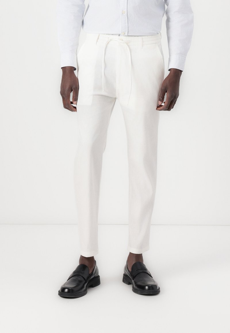 Pantalons blancs à cordon de serrage en tissu léger, avec une coupe fuselée et une texture lisse. Portés avec des mocassins noirs.