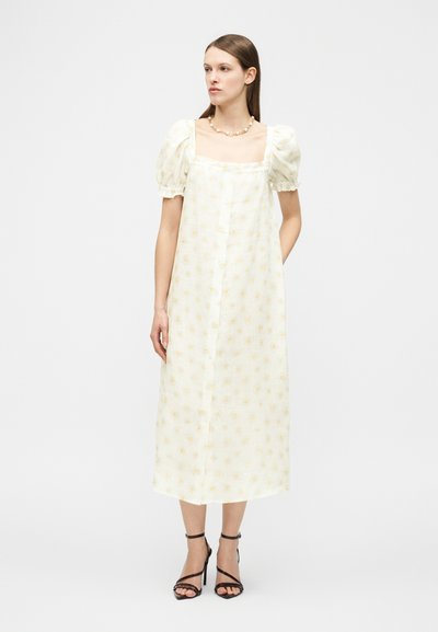 Γυναίκα που φοράει λευκό midi φόρεμα με φουσκωτά μανίκια και κίτρινο λουλουδάτο μοτίβο, συνδυασμένο με μαύρα σανδάλια με λουράκια και ψηλό τακούνι, καθώς και ένα κολιέ.