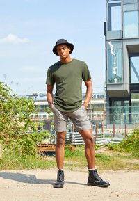 Olijfgroene katoenen t-shirt, grijze geruite shorts, zwarte veterschoenen en een zwarte bucket hat. Buitensetting met moderne gebouwen op de achtergrond.