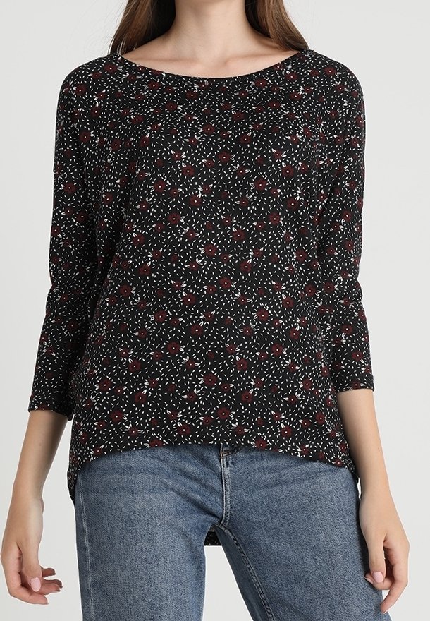 Femme portant un haut noir à manches longues avec de petites fleurs rouges et des taches blanches, associé à un jean bleu en denim.
