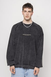 Sudadera negra, de gran tamaño, en tejido suave y texturizado con cuello redondo, que presenta la frase "EXISTENCIA TIMLESS" estampada en dorado en el frente.