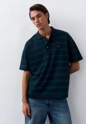 Young man with dark hair wearing a navy blue and green striped polo shirt and blue jeans against a plain white background.