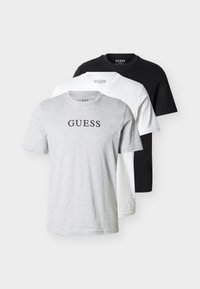 Τρία απλά T-shirts με στρογγυλή λαιμόκοψη σε γκρι, λευκό και μαύρο χρώμα, με το λογότυπο "GUESS" στο κέντρο του στήθους της γκρι μπλούζας.