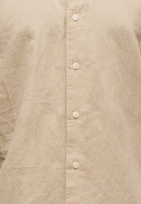 Bež lanena srajca spredaj, ki prikazuje štiri bele gumbe na odprtem robu z vidno teksturo tkanine in rahlimi gubami.