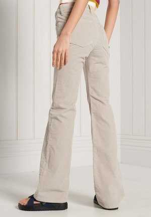 Pantalones de pana beige con corte acampanado y acabado texturado, con dos bolsillos traseros y una cintura de altura media. Combinados con sandalias oscuras.