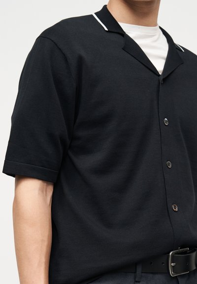 Homme portant une chemise noire à manches courtes avec des boutons et un liseré blanc sur le col, par-dessus un maillot blanc, avec une ceinture noire visible à la taille.