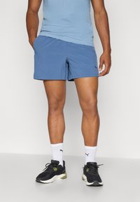 Pantaloni corti sportivi da uomo blu, tessuto leggero, vita elasticizzata, tasche laterali, abbinati a sneakers nere e verdi, calzini bianchi alla caviglia.