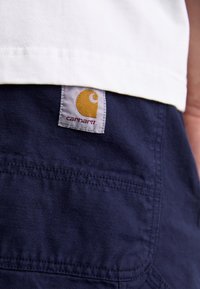 Šortky z tmavě modré látky s bílou štítkem obsahujícím žluté logo a název značky. Na látce jsou viditelné švy a detaily kapes.