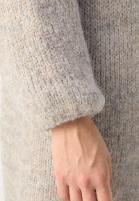Knitted beige sweater sleeve, showing soft, textured yarn with a loose cuff. The hand has visible veins against a light background.