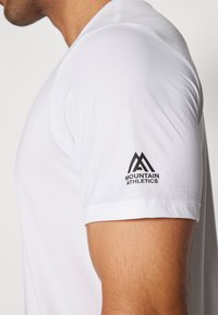 Balta sportinė marškinė su trumpomis rankovėmis, tekstūruota medžiaga ir juodu "Mountain Athletics" logotipu kairiajame petyje.