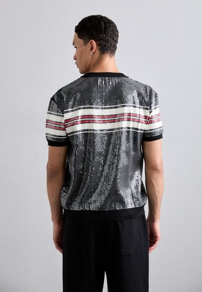 Chemise noire à sequins à manches courtes avec des rayures horizontales blanches et rouges, dotée d'un col et d'un ourlet côtelés, portée sur un pantalon noir.