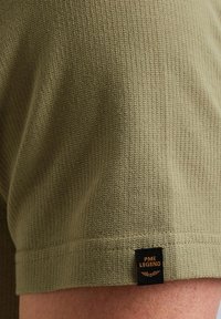 Shorts en tissu côtelé vert olive avec une surface texturée. Présentent une étiquette noire avec "PME LEGEND" en lettres orange au bas.