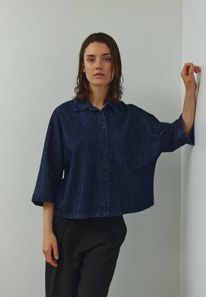 Tmavomodrá rifľová košeľa s golierom, zapínaním na gombíky vpredu a jedným náprsným vreckom. Má voľné, skrátené rukávy a rovný spodný lem.