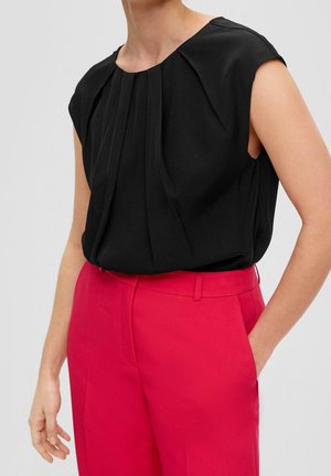 Blouse noire sans manches avec plis à l’avant associée à un pantalon sur mesure rose vif taille haute, main dans la poche.
