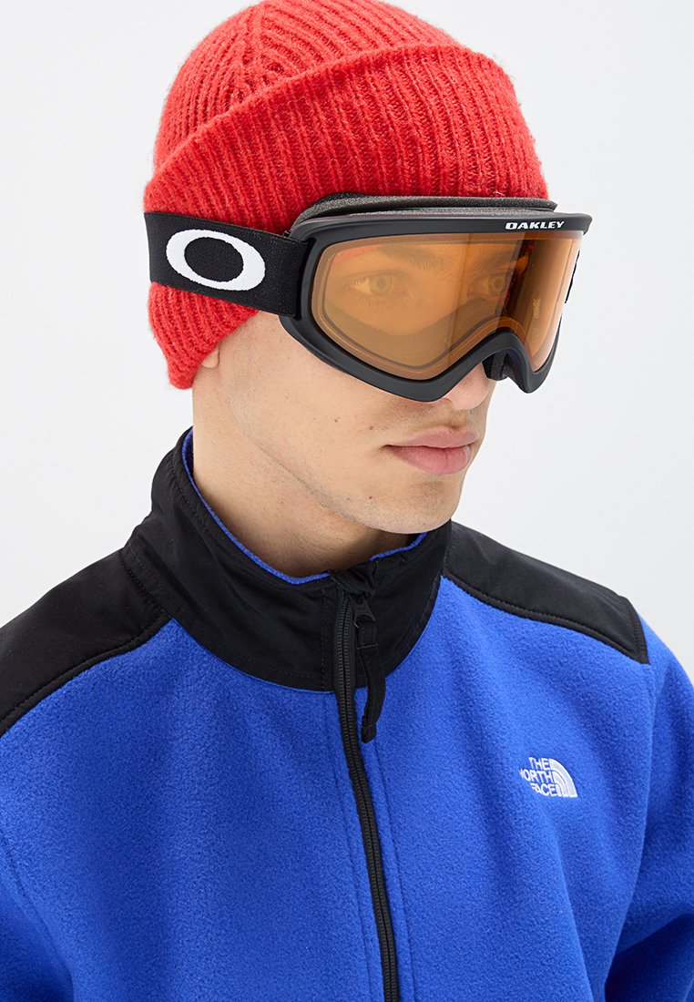 Skibril met oranje lens, zwart frame en witte branding. Draag met een rode gebreide muts en een blauwe fleecjas met zwarte accenten.