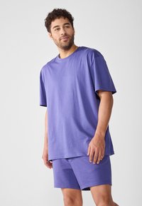 Camiseta de algodón morada con mangas cortas y cuello redondo, combinada con pantalones cortos a juego. Textura suave, corte relajado, diseño sencillo.
