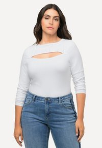 Weißer Longsleeve-Bodysuit mit einem Ausschnitt am Brustbereich, kombiniert mit hoch-taillierten blauen Jeans mit seitlichen Akzenten.