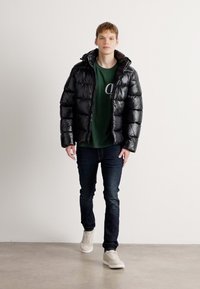 Μαύρο πούφερ μπουφάν με γυαλιστερή φινέτσα, κουκούλα και ελαστικούς καρπούς, συνδυασμένο με πράσινη μπλούζα, σκούρα τζιν και ανοιχτόχρωμα αθλητικά παπούτσια.