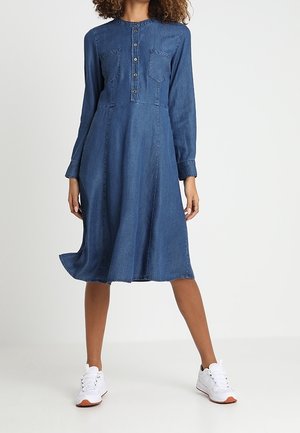 Langærmet kjole i denim med knaplukning, to frontlommer, A-formet silhuet og rå kant. Kombineret med hvide sneakers.