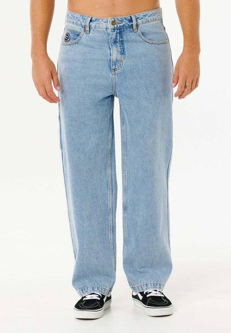 Osoba nosząca luźne jasnoniebieskie dżinsy i czarno-białe tenisówki, stojąca z rękami swobodnie opuszczonymi wzdłuż ciała.