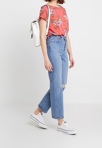 Floral kurzärmliges Koralltop, hellblaue hochgeschnittene Jeans mit Rissdetails und weiße Sneakers. Minimalistischer weißer Rucksack.