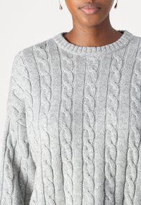 Pull gris en maille avec motif torsadé et encolure ronde côtelée. Texture douce, coupe décontractée et manches légèrement oversize.