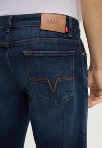 Dunkelblaue Denim-Jeans sind mit einem Leder-Label, kontrastierenden Nähten und einem markanten Design der Gesäßtasche mit einer V-förmigen Naht ausgestattet.