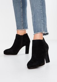 Bottines en suède noir avec un bout arrondi, un haut talon carré et une fermeture éclair sur le côté, associées à un jean bleu à l'ourlet effiloché.