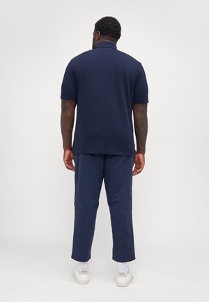 Mand står med ryggen til, iført en marineblå polo, matchende marineblå bukser og hvide Veja-sneakers mod en enkel lys baggrund.