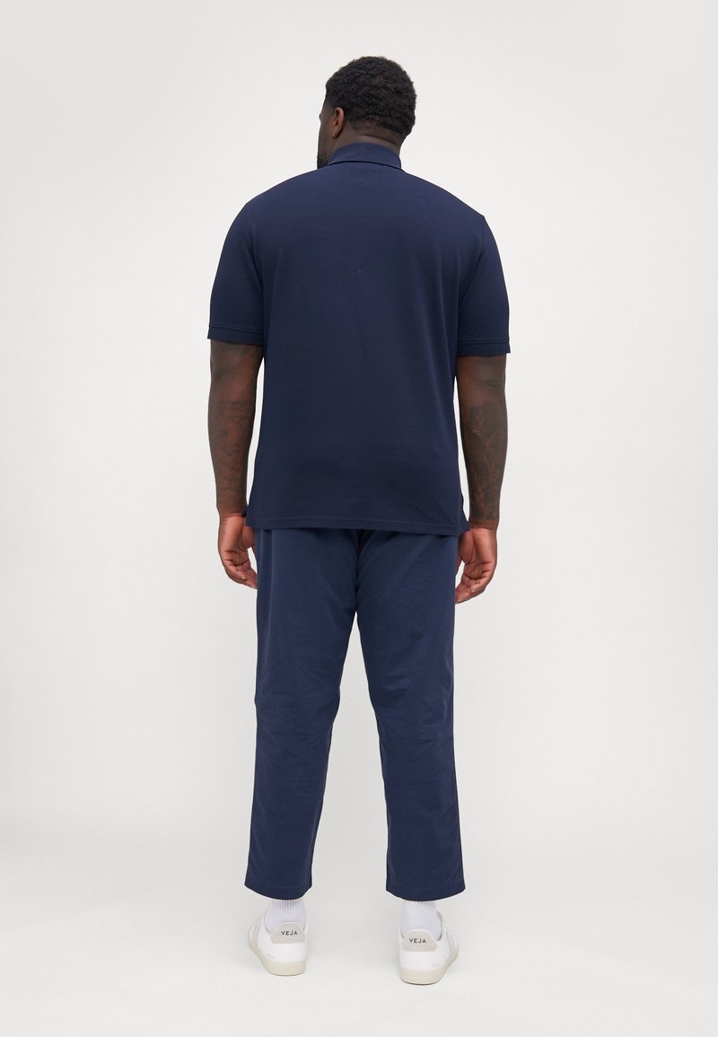 Homme debout, de dos, portant un polo bleu marine, un pantalon assorti et des baskets blanches Veja, sur un fond clair uni.