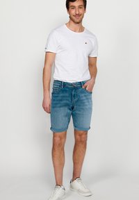 Lahka bela majica s kratkimi rokavi, v kombinaciji z svetloblue denim kratkimi hlačami. Model nosi bele superge in stoji pred nevtralno ozadje.