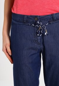 Námořnické denimové kalhoty s pasem na šňůrku, která je vyrobena z tkaného modro-bílého lana. Obsahují přední zapínání na knoflík a volný střih.