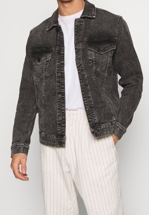 Homme portant une veste en jean noire délavée par-dessus un t-shirt blanc et un pantalon blanc à fines rayures verticales.