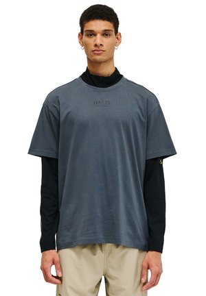 Grå t-shirt med korte ærmer og afslappet pasform, der har et logo på brystet, lagdelt over en sort langærmet skjorte med mesh-struktur.