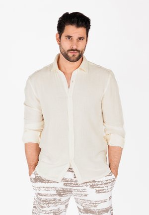 Camisa beige claro de botones con una tela texturizada, mangas arremangadas, combinada con unos pantalones blancos y grises con estampado.