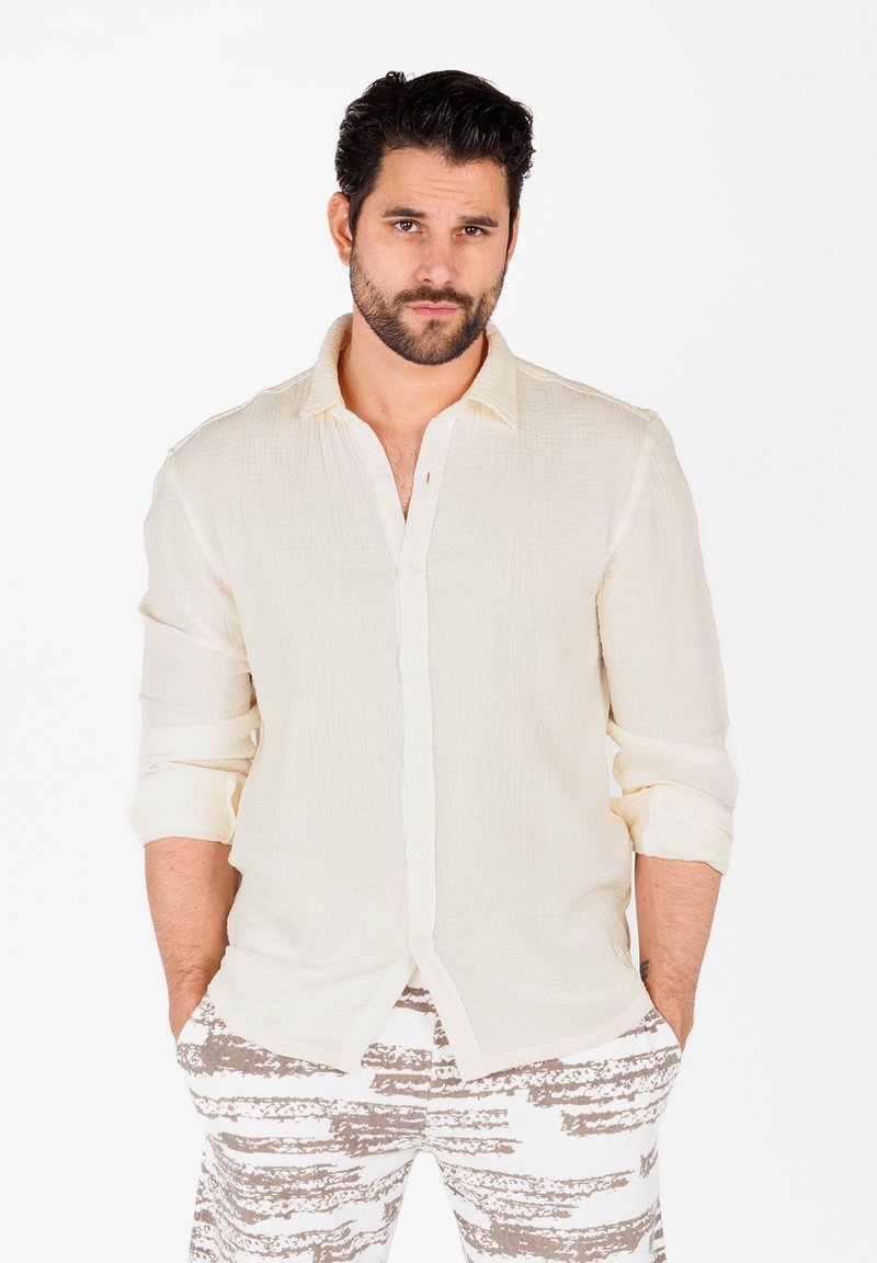 Camisa beige claro de botones con una tela texturizada, mangas arremangadas, combinada con unos pantalones blancos y grises con estampado.