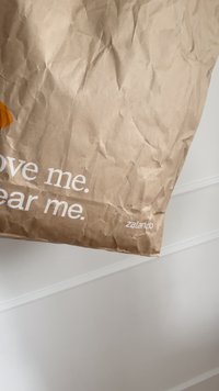 La borsa da shopping in carta kraft presenta una texture spiegazzata, accenti arancioni e testo stampato: "Amami. Indossami." con il logo "zalando" in basso.