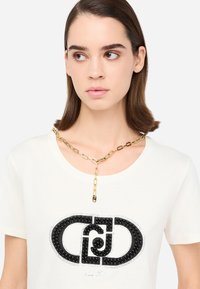T-shirt bianco con un design del logo fatto di perline nere, arricchito da una collana in metallo dorato che evidenzia un distintivo accento a forma di Y.